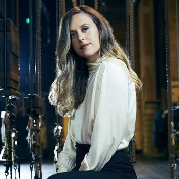 A woman in a white silk blouse sits backstage at a large, historic theater. She is surrounded by stage ropes and pulleys and has light brown, wavy hair around her shoulders.