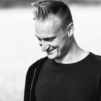 Black and white mid-shot of a smiling Caucasian man with blonde hair cut short at the sides and styled in a quiff at the top wearing a black t-shirt. He stands in a 45 degree left side profile with his head slightly angled down towards the floor.