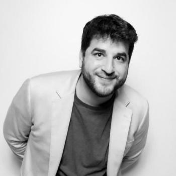 director, Zach Wolff in a suit jacket and shirt smiles at the camera in black and white